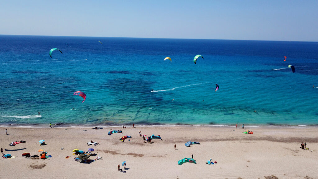 Vibrant view from a hill of a lively beach scene with people sunbathing and kite surfing, illustrating the dynamic lifestyle possible with Rural Land for Sale in Lefkada.