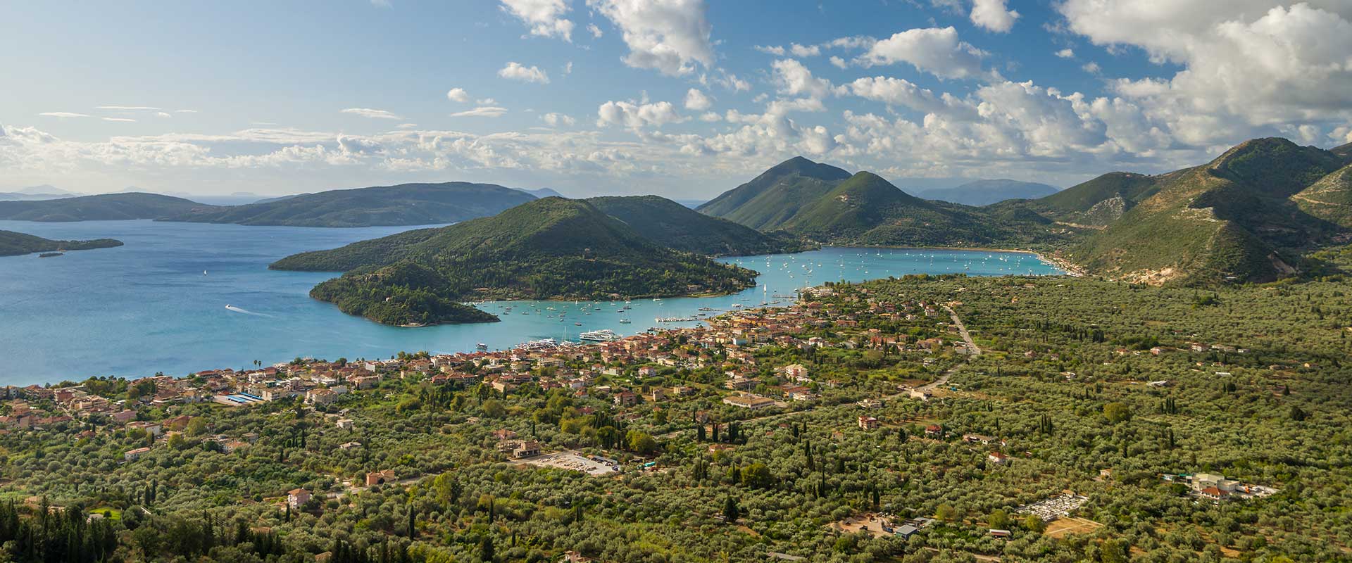 An aerial view captured by a drone showcasing the beautiful town of Nidri in Lefkada, Greece. The image highlights the picturesque landscape with lush greenery, crystal-clear turquoise waters, and charming buildings. In the foreground, there is a prominent sign reading 'Land for Sale in Nidri, Lefkada' indicating available property opportunities amidst this stunning location.